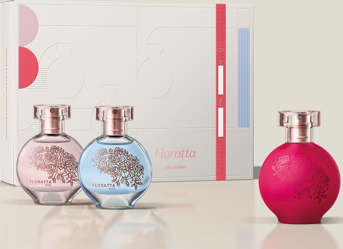 A imagem mostra uma linha de produtos de perfumaria da marca Floratta. Há três frascos de perfume em vidro transparente com detalhes gravados em rosa e azul claro, juntamente com uma caixa de papelão branca com elementos gráficos em rosa, vermelho e azul.
