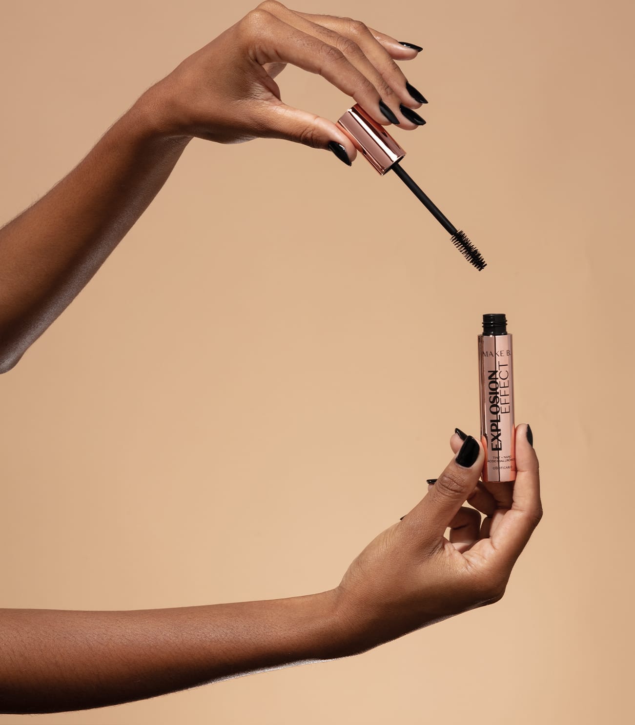 Uma mão feminina segura um tubo rosa furta-cor com letras pretas de máscara de cílios Make B. Explosion Effect e a outra mão segura a tampa rosa furta-cor com haste e aplicador pretos, sobre fundo em tons de bege.