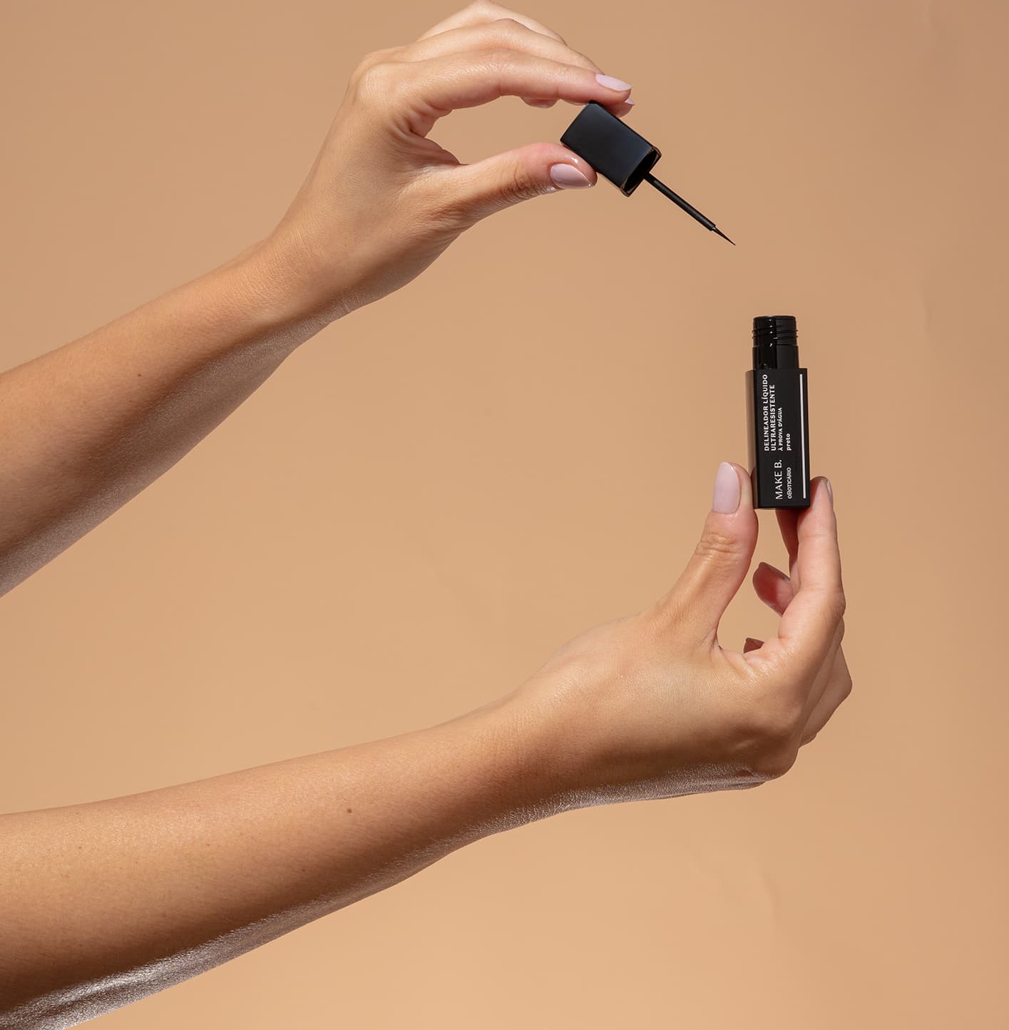 Uma mão feminina segura um tubo preto  com letras brancas de delineador líquido para olhos na cor preta da marca Make B., e a outra mão segura a tampa com haste e aplicador fino, tudo sobre fundo bege.
