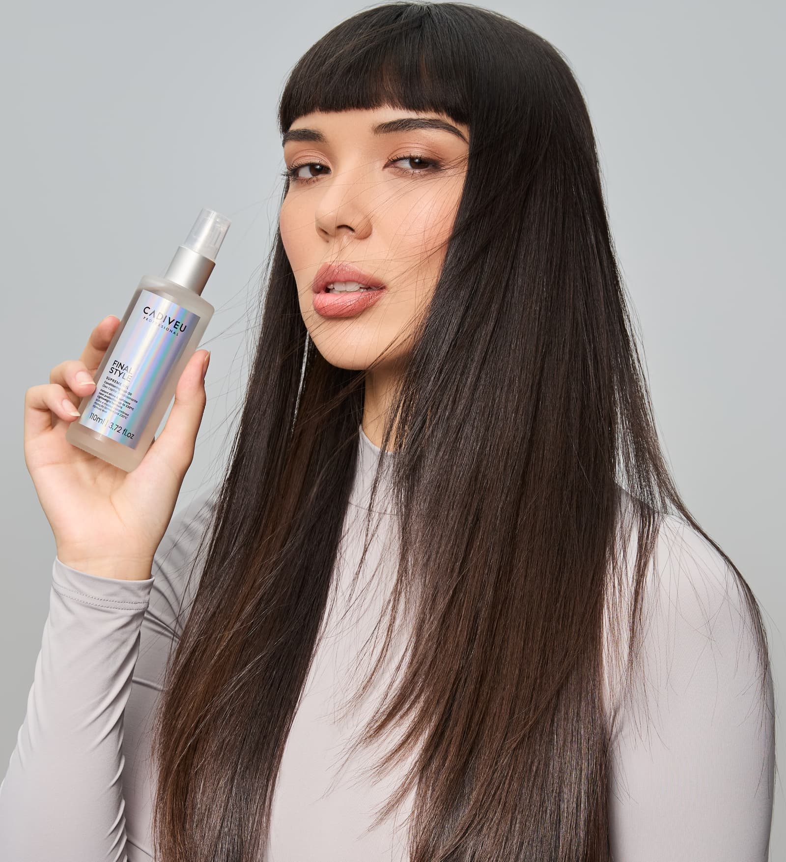 A imagem mostra uma modelo com cabelos lisos, longos e escuros, segurando o Óleo Capilar Cadiveu Professional Final Style Supreme Oil. Ela usa uma blusa cinza claro com manga longa.