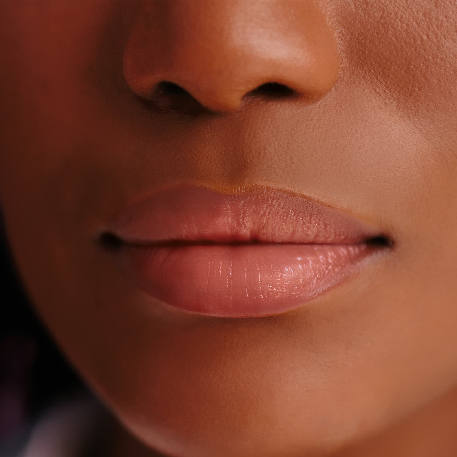 A imagem mostra um close-up dos lábios de uma pessoa, com uma textura suave e brilhante. A cor dos lábios é um rosa claro e natural, com um acabamento levemente acetinado. Não é possível identificar a pessoa.