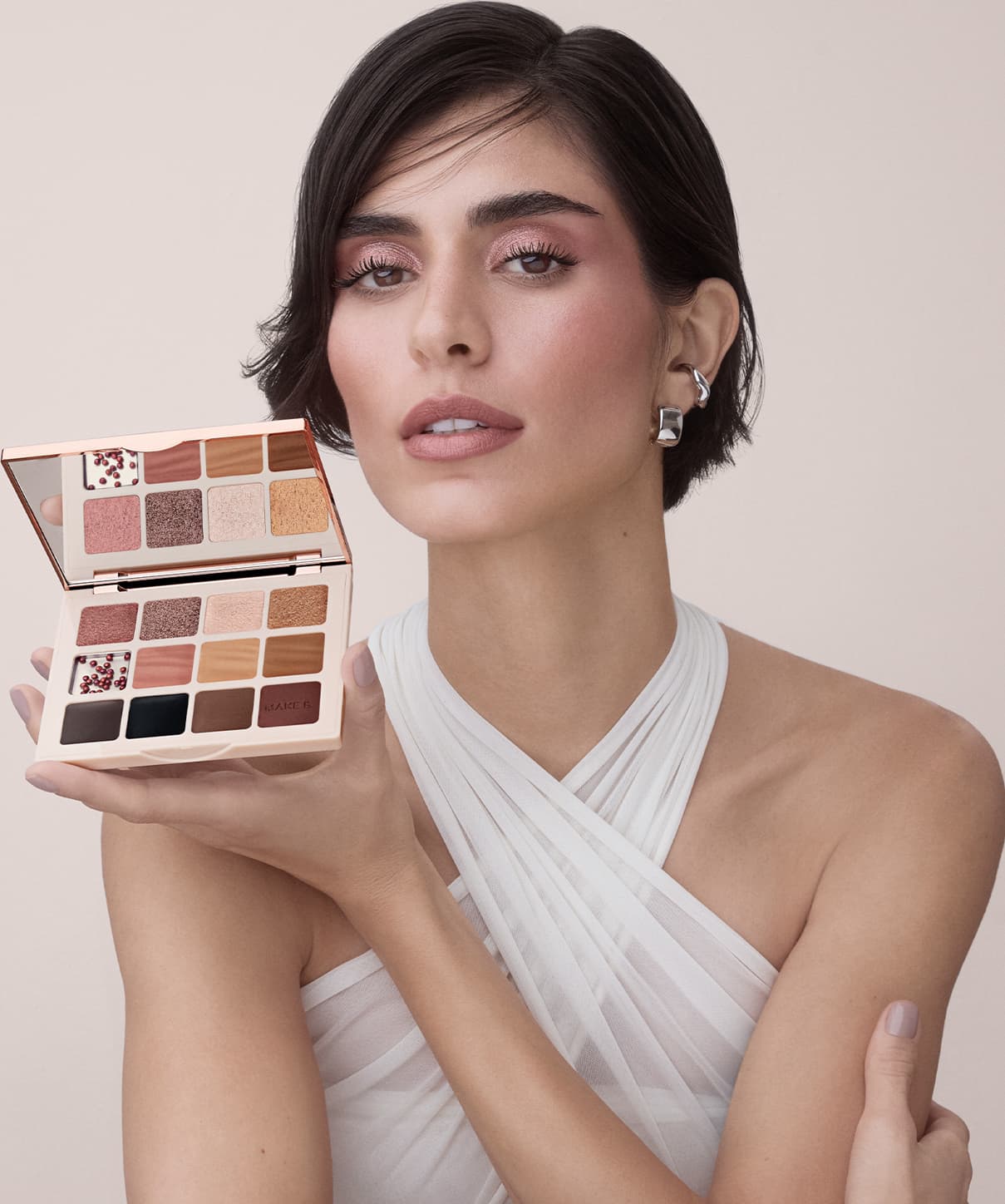 A paleta de sombras de maquiagem da marca Make B. A embalagem é retangular, com um design elegante em tons de rosa e dourado. A paleta está sendo segurada por uma modelo com cabelos escuros e maquiagem suave.