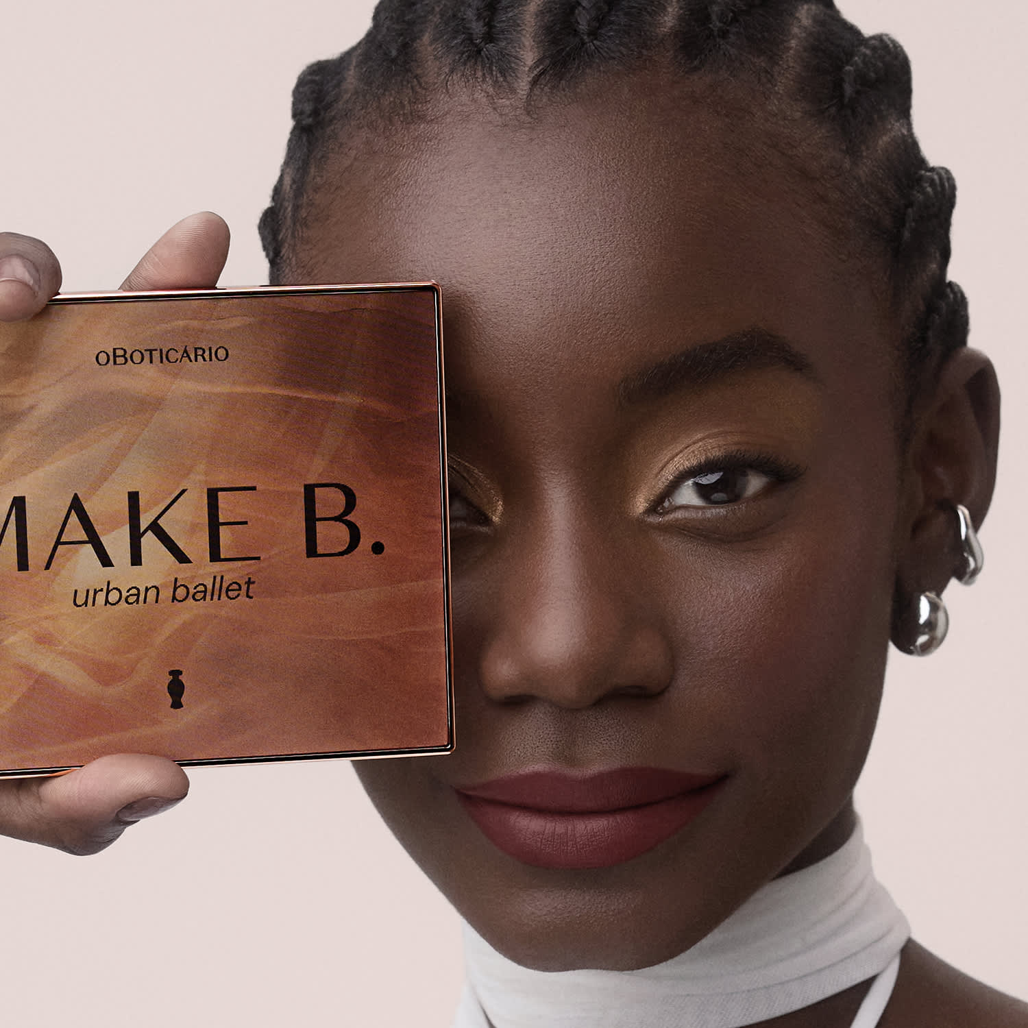 A caixa de maquiagem da marca Make B. urban ballet é exibida na imagem. A caixa é de cor marrom e possui o nome da marca e o tipo de produto impresso em letras grandes. A pessoa segura a caixa próxima ao rosto, permitindo uma visão clara da embalagem.