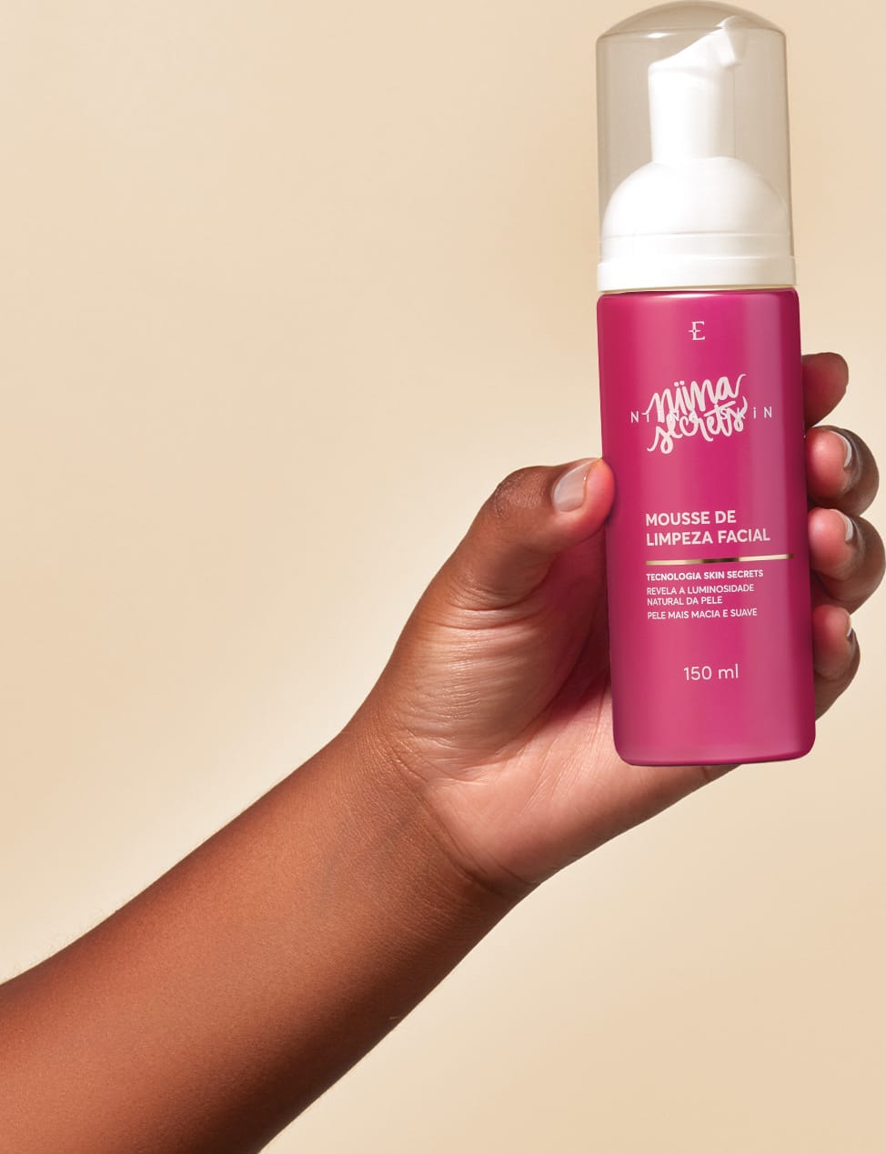 Produto de limpeza facial da marca Niina Secrets, em embalagem rosa, sendo segurado por uma mão em ambiente neutro de fundo bege. O produto é apresentado como uma mousse de limpeza facial com tecnologia skin secrets.