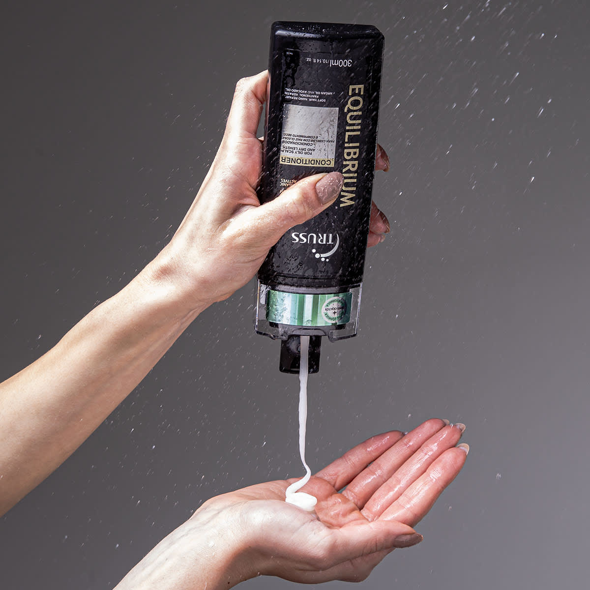 A imagem mostra uma mão segurando um frasco de condicionador da marca Truss, linha Equilibrium. O frasco está molhado, aparentemente sendo usado no chuveiro ou banheiro. Também é possível ver o seu conteúdo, que é cremoso e branco.