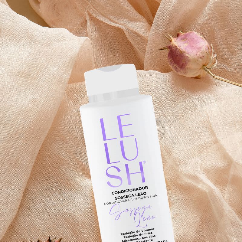A imagem mostra um frasco de condicionador branco com o nome da linha escrito em letras roxas. A composição também inclui um tecido bege delicado e um botão de rosa rosada, criando uma atmosfera suave e natural.