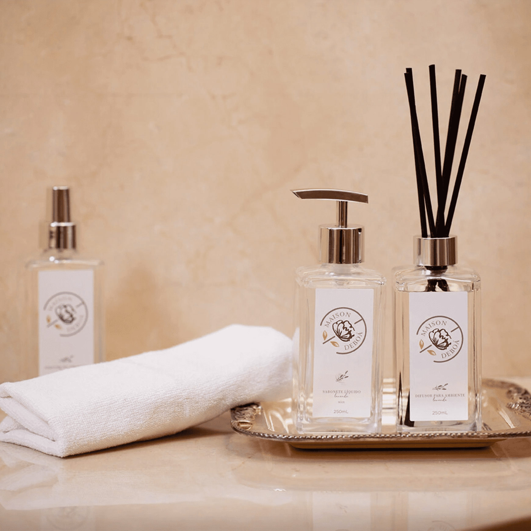 Produtos de fragrância e cuidados corporais da marca Maison Deboá. 3 frascos de vidro transparente com tampas douradas.. Um frasco é um pulverizador, outro tem um dosador de bomba e o terceiro contém difusores de aromas. Também há uma toalha branca