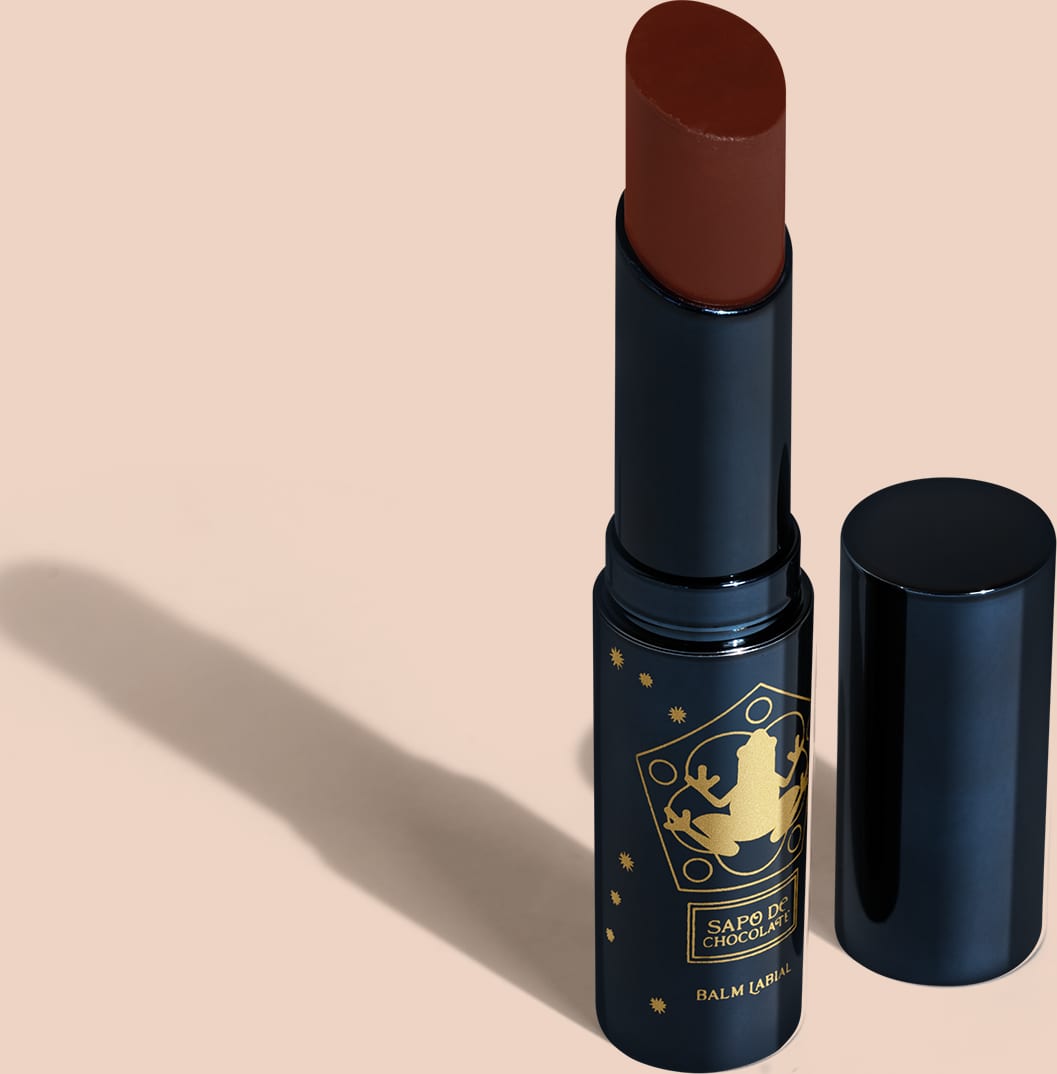 Balm em formato de bastão com uma cor marrom. A embalagem é um tubo azul escuro com detalhes dourados que incluem um símbolo de um caldeirão. O produto tem uma aparência elegante e sofisticada, com o tubo azul fosco contrastando com os detalhes dourados