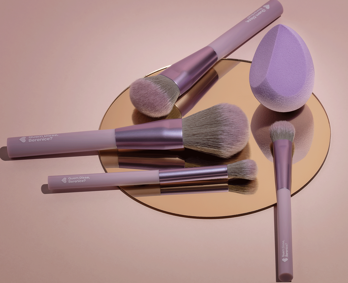 A imagem mostra uma variedade de pincéis de maquiagem da marca Quem Disse, Berenice?. Os produtos têm cabos longos e finos em tons de roxo e marrom, com cerdas macias nas pontas. Há pincéis de diferentes formatos e tamanhos e uma esponja roxa.
