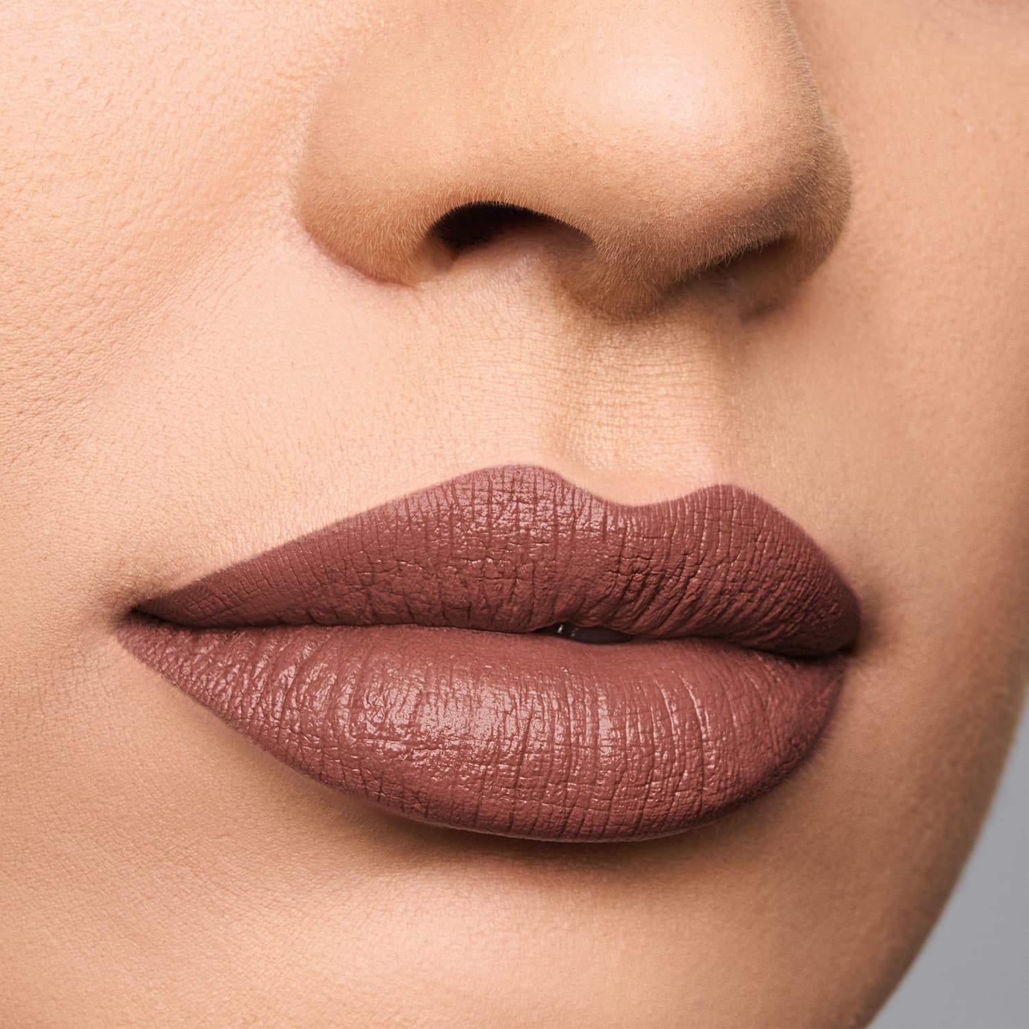 A imagem mostra um close-up dos lábios de uma pessoa, com uma maquiagem de batom da marca Quem Disse, Berenice?. O batom tem uma tonalidade marrom-acinzentada, com uma textura cremosa e brilhante. Os lábios estão bem definidos.