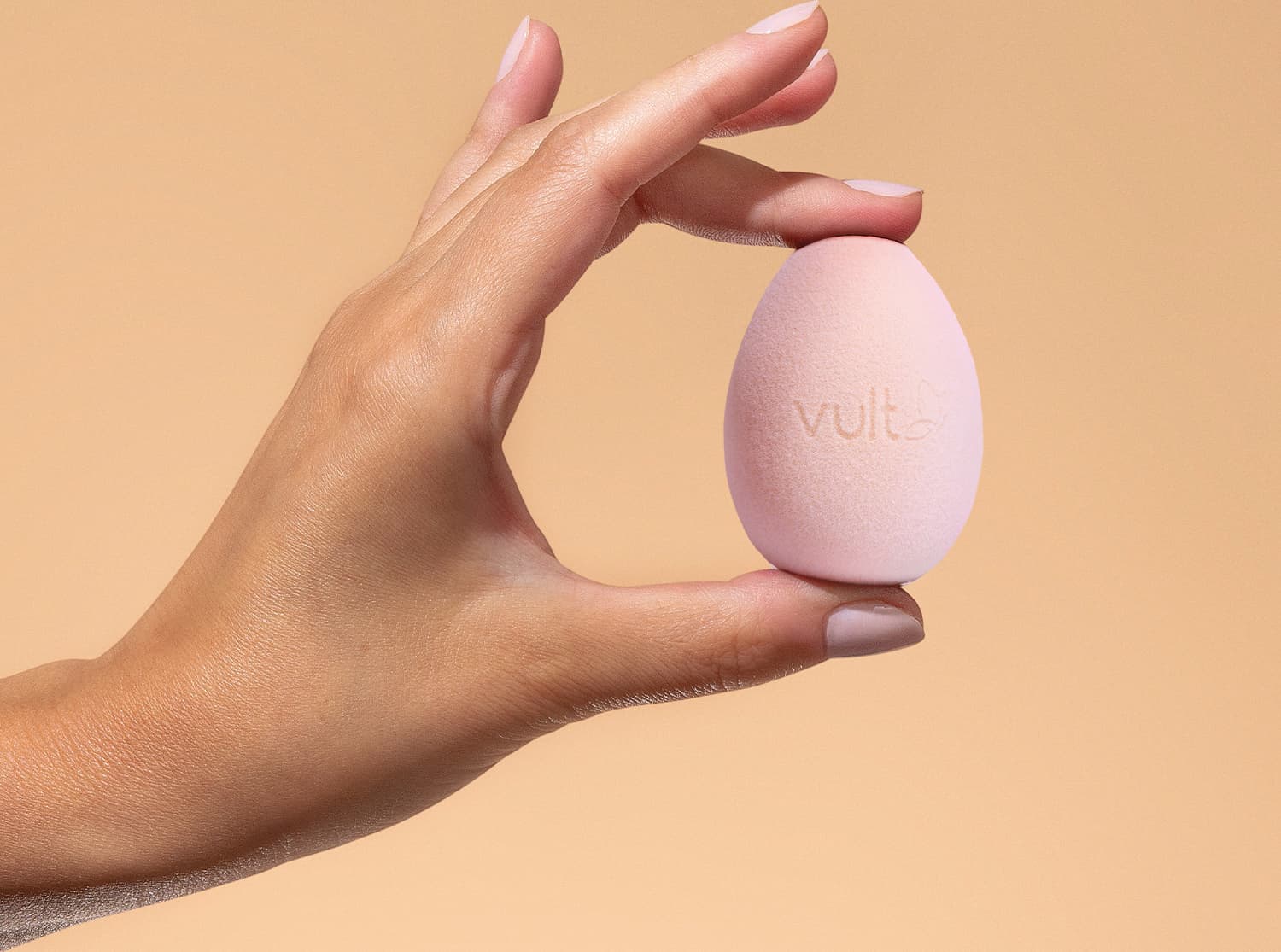 Mãos segurando uma esponja de maquiagem Vult em um fundo laranja, sugerindo um ambiente de cuidados com a pele e produtos de beleza.