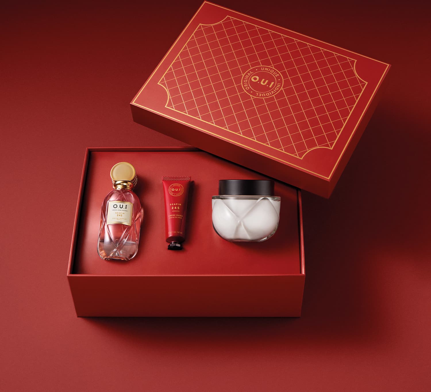 Itens estão dispostos dentro da caixa vermelha de presente quadrada com a logo da marca O.U.i estampada em dourado. A caixa possui um padrão de grade dourado sobre o fundo vermelho. Os produtos estão encaixados em seus respectivos lugares dentro da caixa.