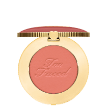 Blush compacto de maquiagem com embalagem redonda dourada. A tampa superior é rosa com a inscrição "Too Faced" em letras cursivas. Um pequeno coração dourado adorna o centro da tampa.
