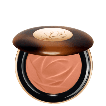 A imagem traz um sérum bronzeador compacto em uma embalagem. A tampa superior é marrom com alguns detalhes mais claros e tons mais escuros. O logotipo da marca aparece gravado em dourado no centro da tampa e o produto possui uma cor bege.