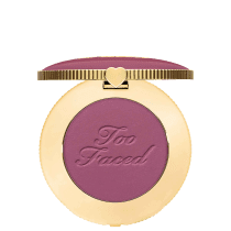 Blush compacto de maquiagem com embalagem redonda dourada. A tampa superior é roxa com a inscrição "Too Faced" em letras cursivas. Um pequeno coração dourado adorna o centro da tampa.
