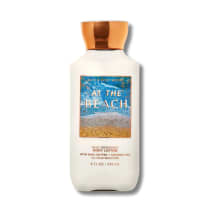 Frasco de loção corporal da Bath & Body Works com rótulo azul e dourado, apresentando a frase "At the Beach" e informações sobre os ingredientes de manteiga de karité e óleo de coco.