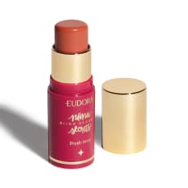 O produto apresentado é um blush stick da linha Niina Secrets da marca Eudora. Possui uma embalagem rosa com detalhes dourados, incluindo o nome da marca e da linha. O produto tem formato cilíndrico e tampa dourada.