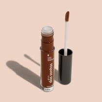 Frasco cilíndrico transparente contendo um gloss labial marrom. Rótulo na embalagem com o nome do produto "Gloss dos Sonhos" e a descrição "Quem disse, berence?". Ao lado, a tampa preta do produto, que acompanha um aplicador de espuma.