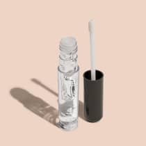 Gloss labial transparente em frasco cilíndrico de vidro com tampa plástica branca. Ao lado, um aplicador de maquiagem em formato de pincel com cabo preto. Embalagem com o texto "gloss dos sonhos" visível. Cores predominantes são transparente e preto.