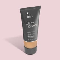 O produto apresentado é uma base líquida da marca Tô no Glow, com embalagem em tubo plástico preto e branco. O rótulo exibe o nome da marca e a descrição "Radiância natural, leveza e conforto". O produto é classificado como "Base líquida".