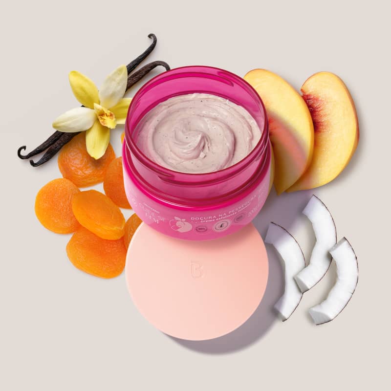 O produto apresentado é um creme esfoliante para a pele, em uma embalagem rosa Ao redor, há diversos ingredientes naturais, como flores de baunilha, fatias de pêssego e rodelas de coco, sugerindo que o produto possui uma fórmula com ingredientes naturais.