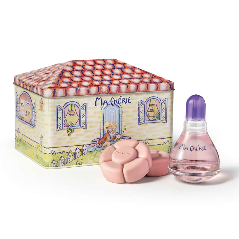 Kit Ma Chérie incluindo uma caixa decorada com ilustrações de uma casa e uma figura feminina, com tampa em formato de telha rosa. Um frasco transparente com líquido rosa e tampa roxa.Também há sabonetes em formato de flores na cor rosa claro.