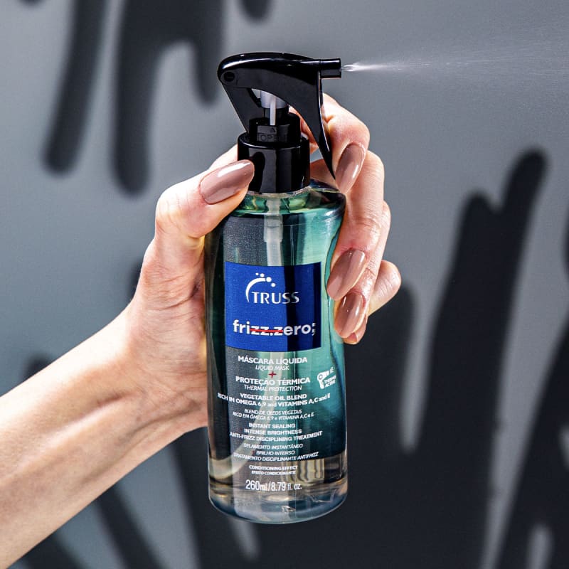 A imagem mostra uma mão segurando um frasco de um produto de cuidados capilares líquido chamado "Frizz Zero" da marca Truss. O frasco é transparente com um líquido azul dentro e detalhes do produto escritos em português e inglês.