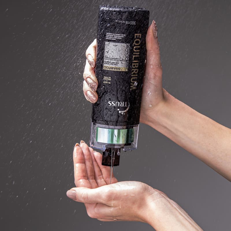 A imagem mostra uma mão segurando um frasco de shampoo da marca Truss, linha Equilibrium. O frasco está molhado, aparentemente sendo usado no chuveiro ou banheiro. Também é possível ver o seu conteúdo, que é cremoso e transparente.