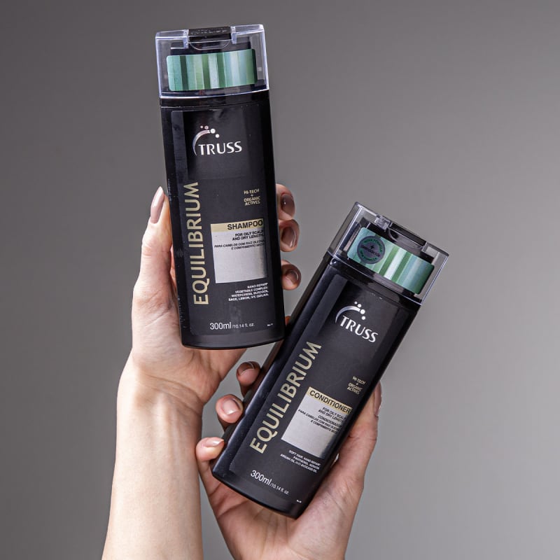A imagem mostra duas embalagens de produtos para cabelos da marca Truss. Um é um shampoo e o outro é um condicionador, da linha Equilibrium. As embalagens são pretas com detalhes em verde e estão sendo seguradas por uma mão.