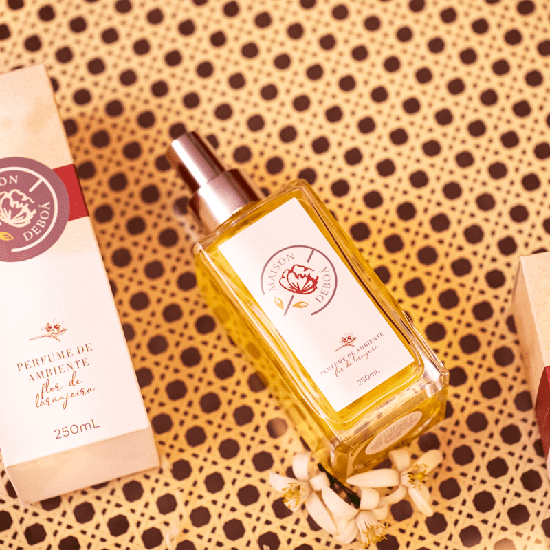 Perfume ambiente em frasco, posicionado sobre um fundo estampado com bolinhas marrons sobre bege. Ao lado, há uma caixa retangular do mesmo produto com detalhes em vermelho e flores secas brancas.