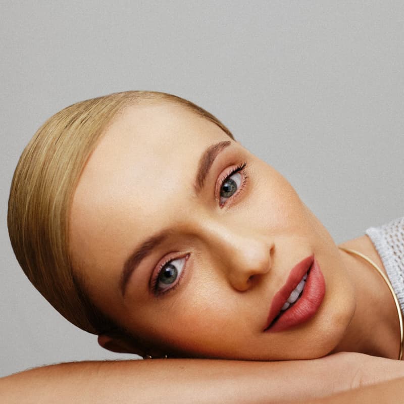 A imagem é um rosto feminino com maquiagem. Observa-se uma pele branca, olhos azuis expressivos, sobrancelhas bem definidas e lábios com um tom de rosa suave. A iluminação e o fundo neutro destacam as características faciais de forma elegante e sutil.