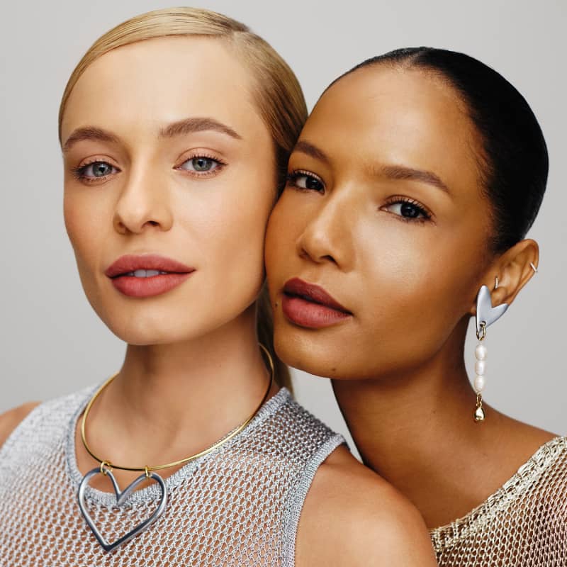 A imagem mostra dois rostos femininos em close-up, com expressões sérias e olhares diretos. Ambas as mulheres usam maquiagem com tons neutros e naturais, destacando seus traços faciais. Elas vestem roupas com detalhes em malha metálica.