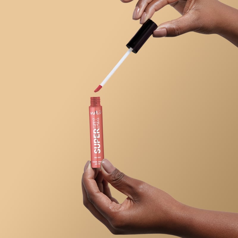 A imagem mostra uma mão segurando o produto aberto do Batom Líquido da marca Vult, na cor castanho. A embalagem do produto é um tubo vermelho com o nome da marca "Vult" impresso.