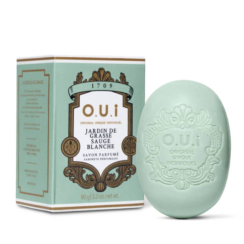 Sabonete perfumado em formato oval com relevo da marca O.U.I. ao lado da embalagem verde-menta com detalhes dourados e rótulo com informações sobre o produto "Jardin de Grasse Sauge Blanche".