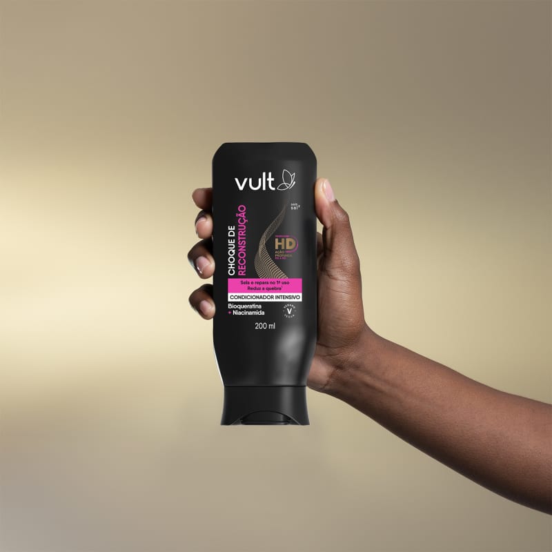 A imagem apresentada é uma mão segurando o produto, o condicionador intensivo da marca Vult. A embalagem é um tubo preto com o logotipo da marca e informações sobre o produto, como "Choque de Reconstrução" e "Tecnologia HD".