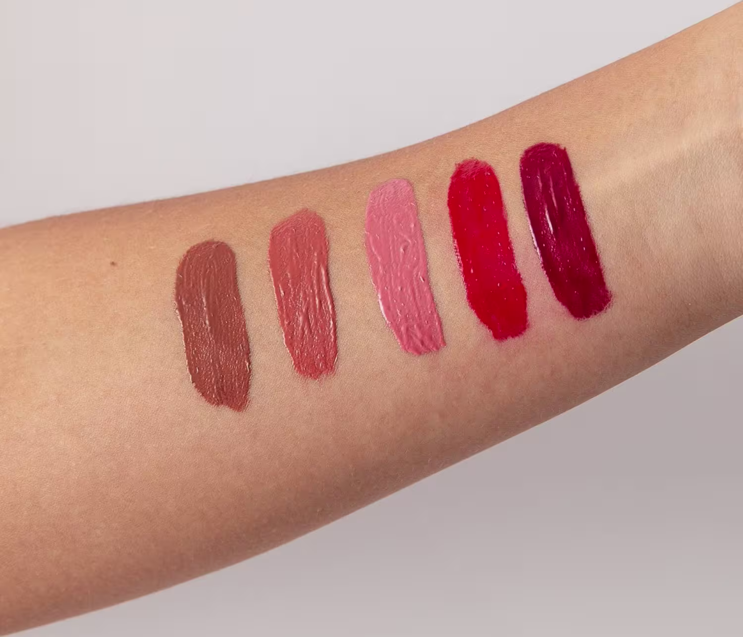 São cinco amostras retangulares aplicadas no antebraço, variando do marrom terroso ao vermelho intenso. As cores vão do marrom avermelhado, passando pelo rosa claro, rosa escuro, vermelho vivo até o vermelho profundo.