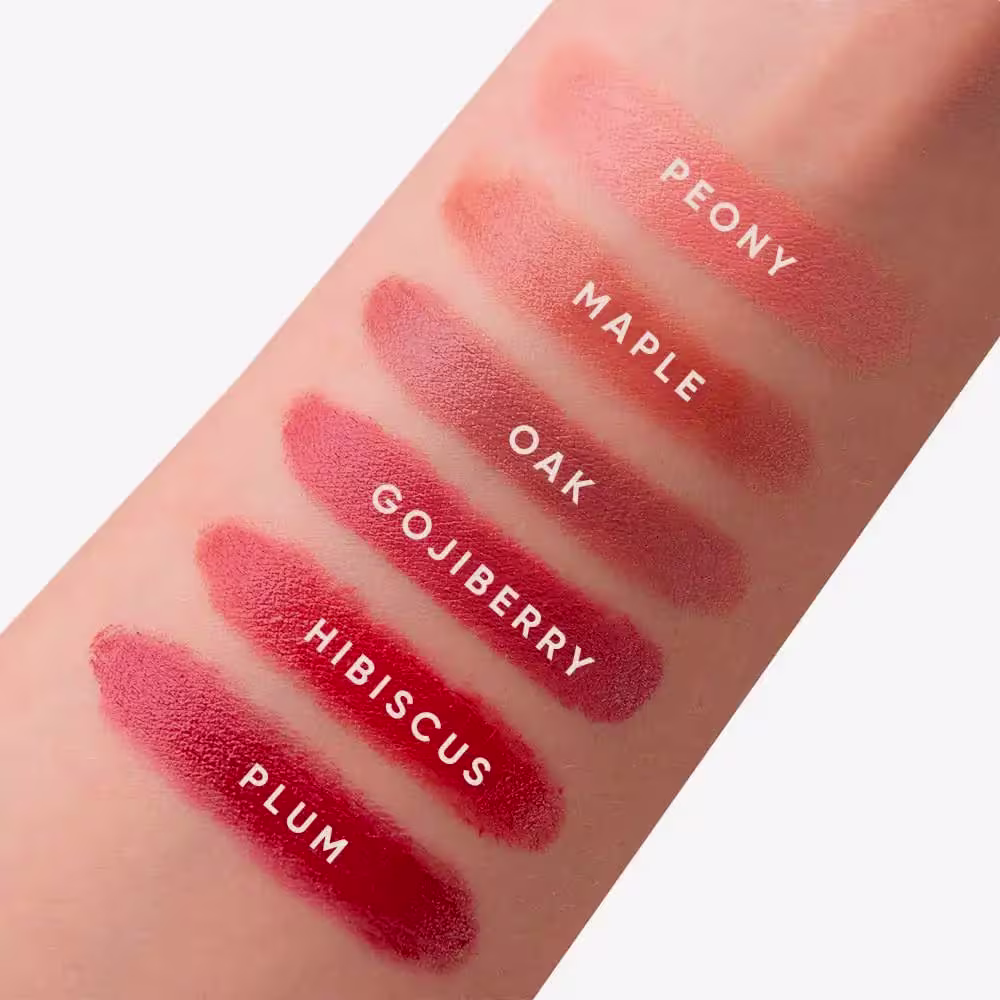 A imagem mostra uma série de amostras da cor do blush em diferentes tons de vermelho, variando de um rosa claro a um vermelho escuro. Os nomes dos tons estão impressos sobre cada amostra, incluindo "Peony", "Maple Oak", "Goji Berry" e "Hibiscus Plum".