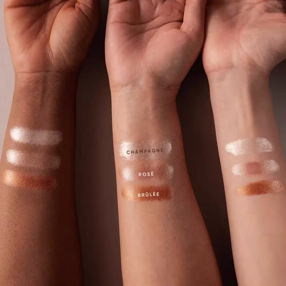A imagem mostra o resultado de cor em diferentes tons de pele.  Há três diferentes tonalidades, com os rótulos "Champagne", "Rosé" e "Brûlée" visíveis nos braços. A apresentação é um swatch, com as diferentes tonalidades aplicadas diretamente na pele.
