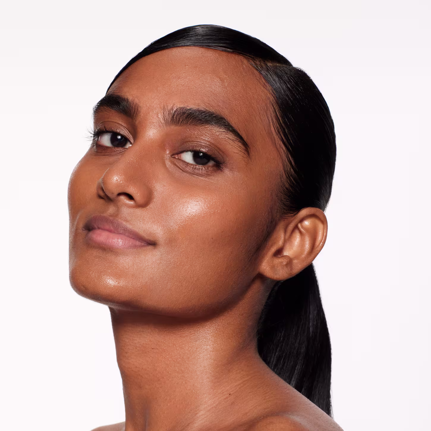 Esta imagem mostra um close-up do rosto de uma mulher jovem com pele negra brilhante e olhos castanhos expressivos. Ela tem sobrancelhas bem delineadas e lábios cheios e brilhantes, sugerindo o uso de produtos de maquiagem de alta qualidade.