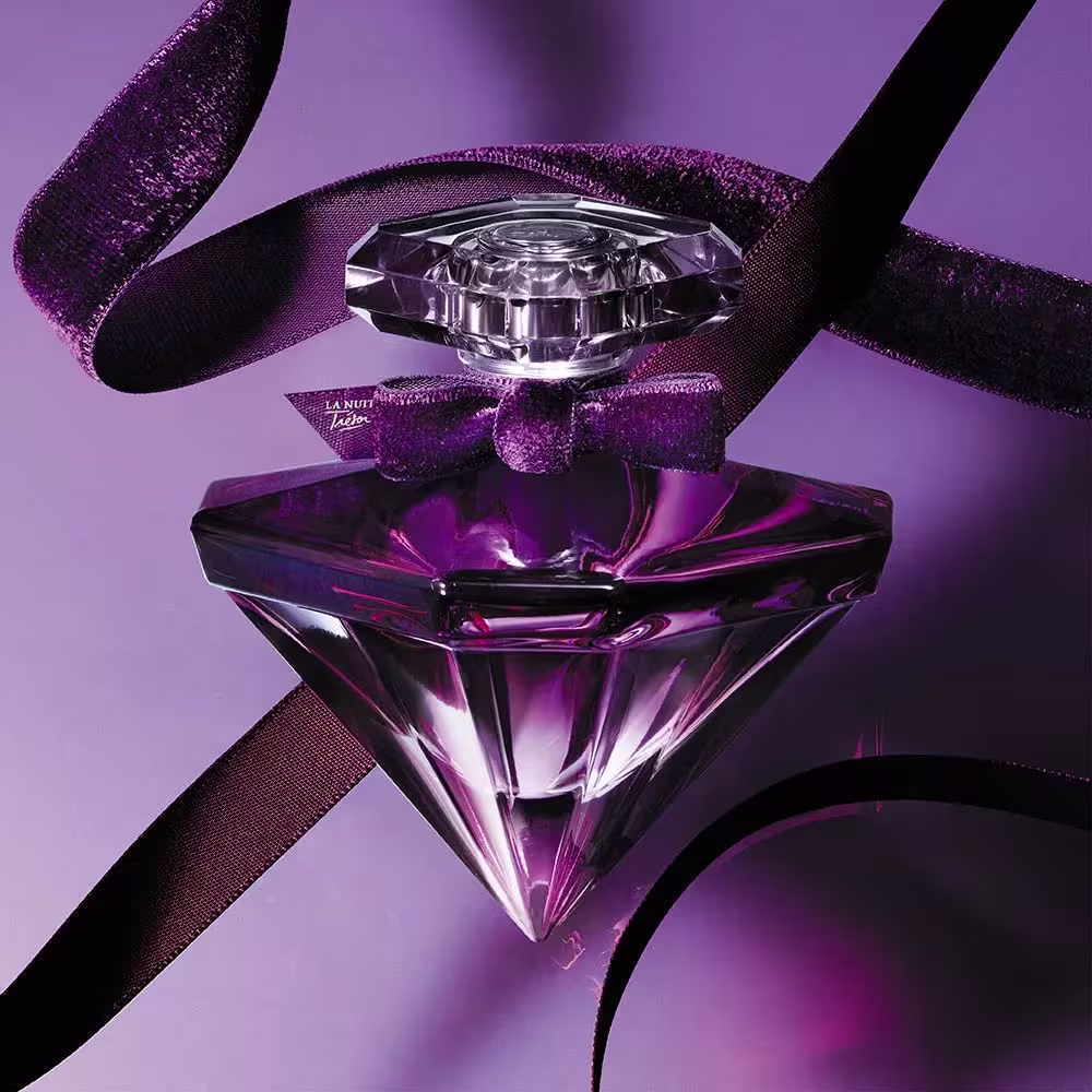 Perfume em frasco de cristal triangular com design facetado. A tampa do frasco também é de cristal facetado. O frasco está envolto por uma fita de cetim roxo brilhante com a inscrição "La Nuit Trésor".