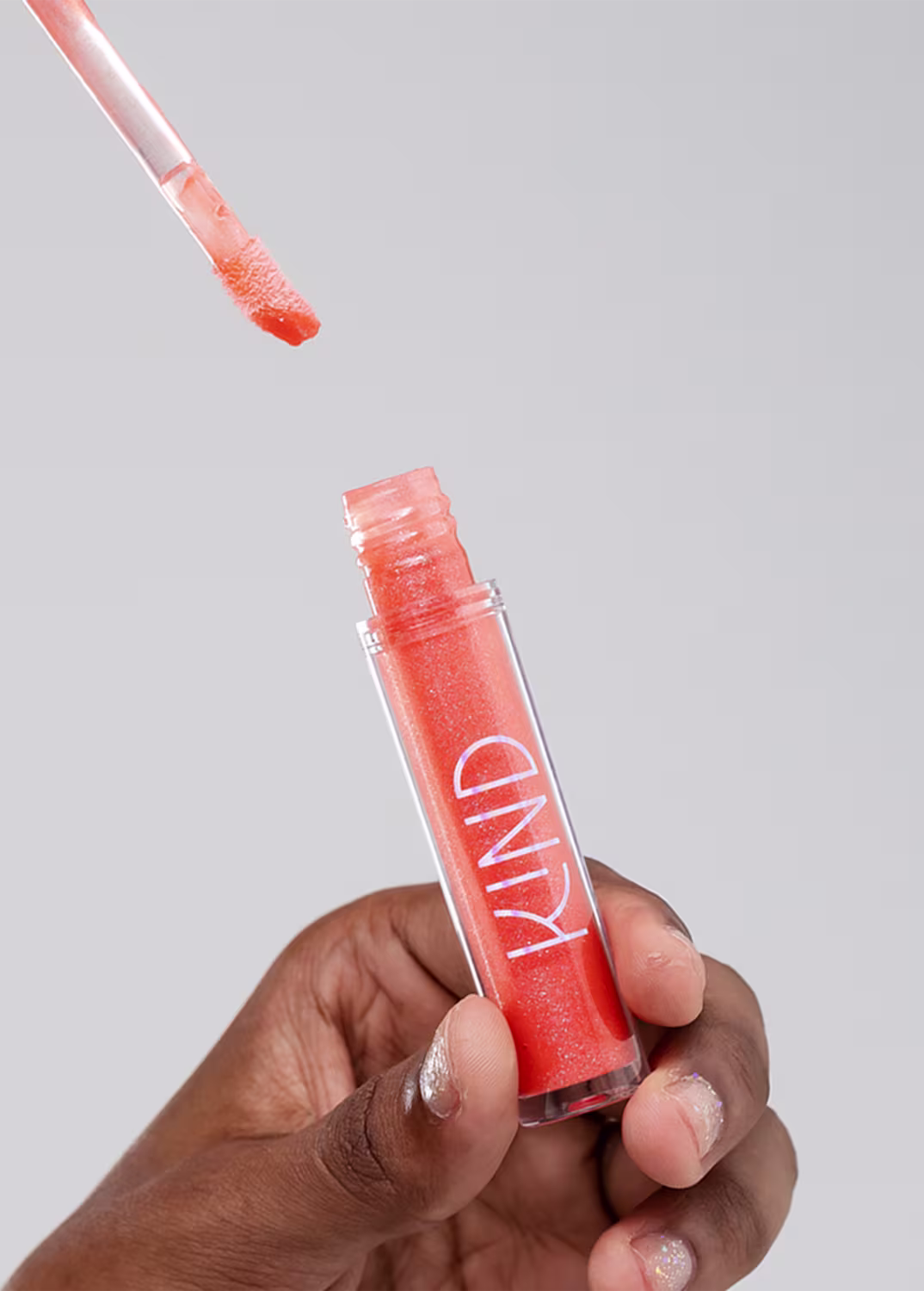 A imagem mostra a embalagem do gloss, aberta, sendo segurado por uma mão negra com unhas curtas.