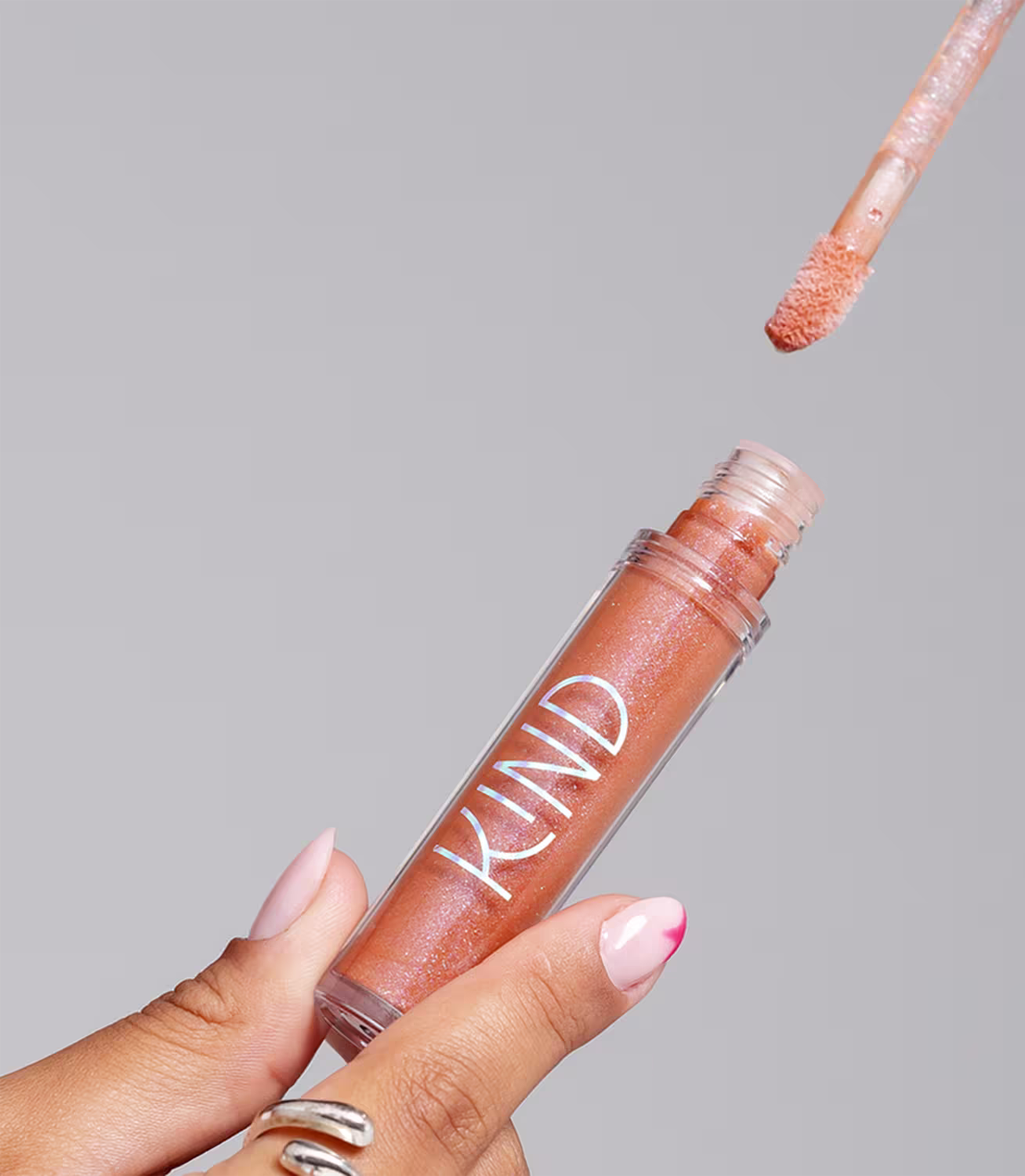 A imagem mostra a embalagem do gloss, aberta, sendo segurado por uma mão com unhas claras e anéis prateados.