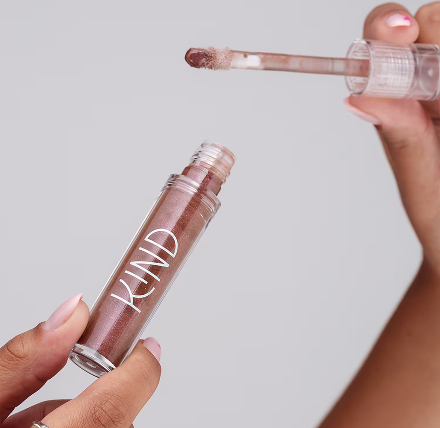 A imagem mostra a embalagem do gloss, aberta, sendo segurado por uma mão com unhas claras e anéis prateados.