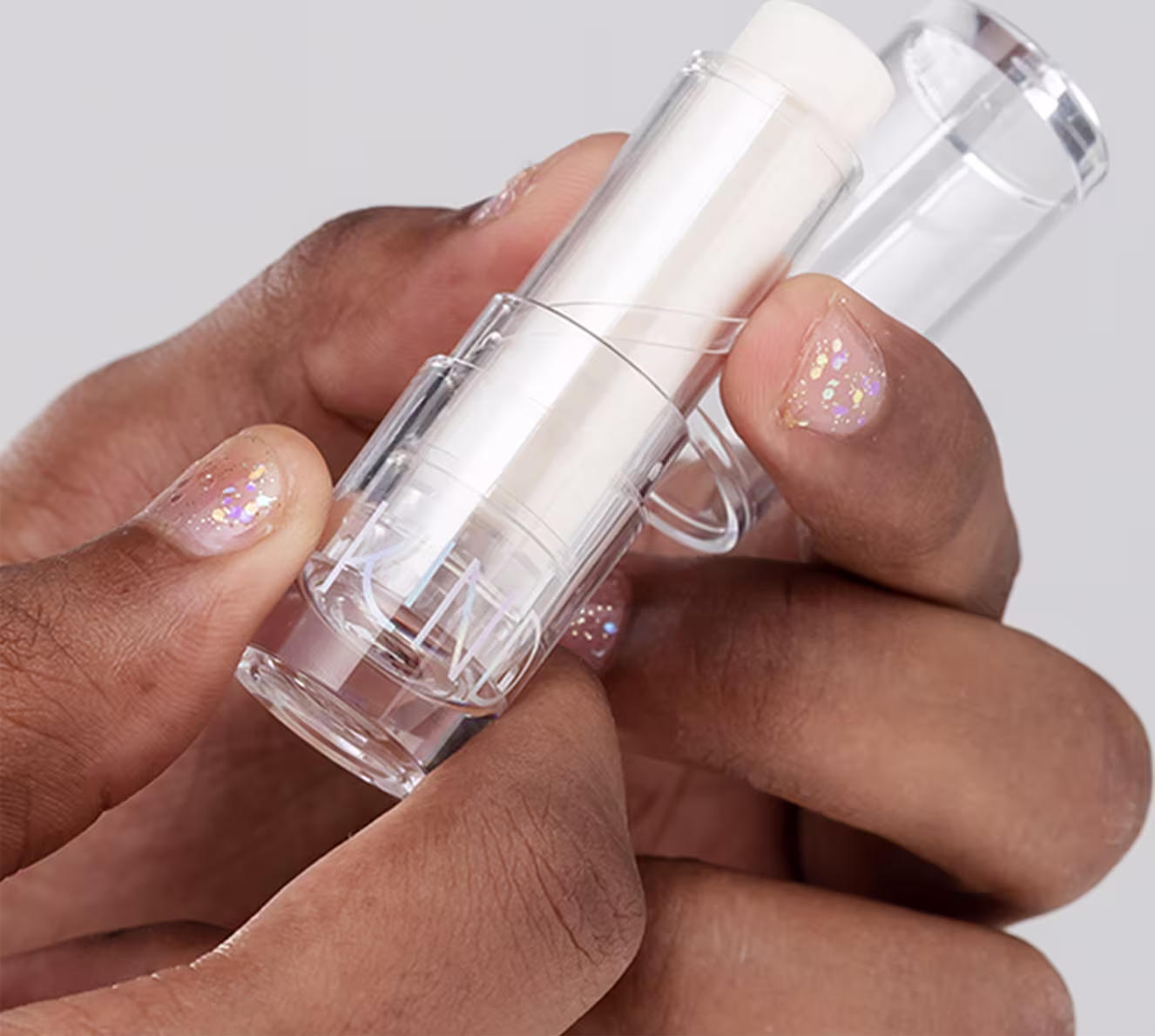 A imagem mostra a embalagem do balm, aberta, sendo segurado por duas mãos negras, com unhas curtas e esmalte com glitter.