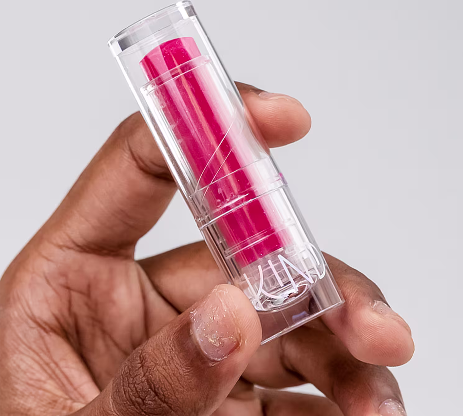 A imagem mostra a embalagem do balm, fechada, sendo segurado por uma mão negra, com unhas curtas e esmalte com glitter.