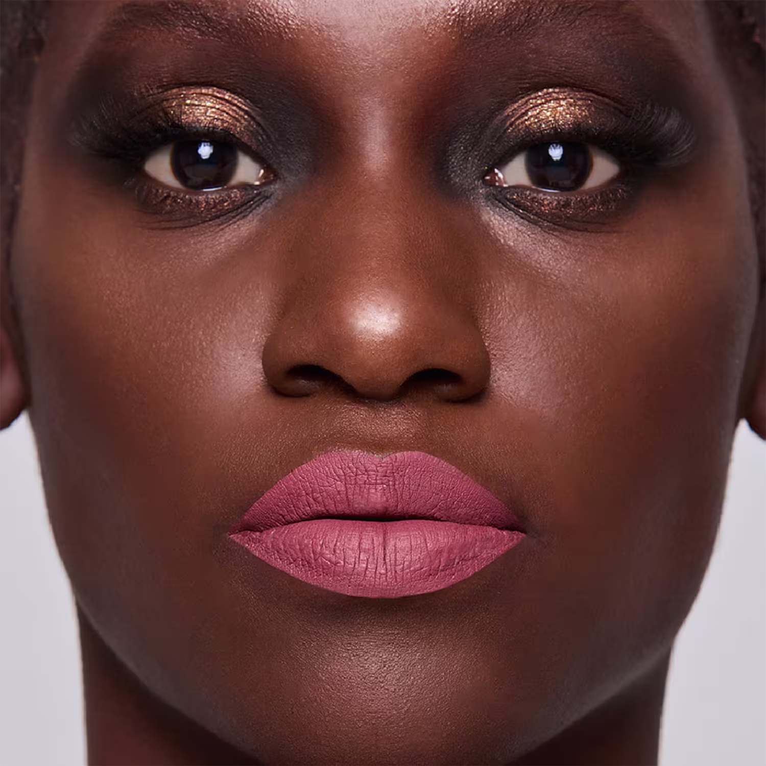 Esta imagem mostra uma mulher negra com maquiagem elaborada. A pele tem um lindo tom marrom vibrante e brilhante. Os lábios foram maquiados com um batom de cor rosa envelhecido, realçando a boca bem delineada e cheia.