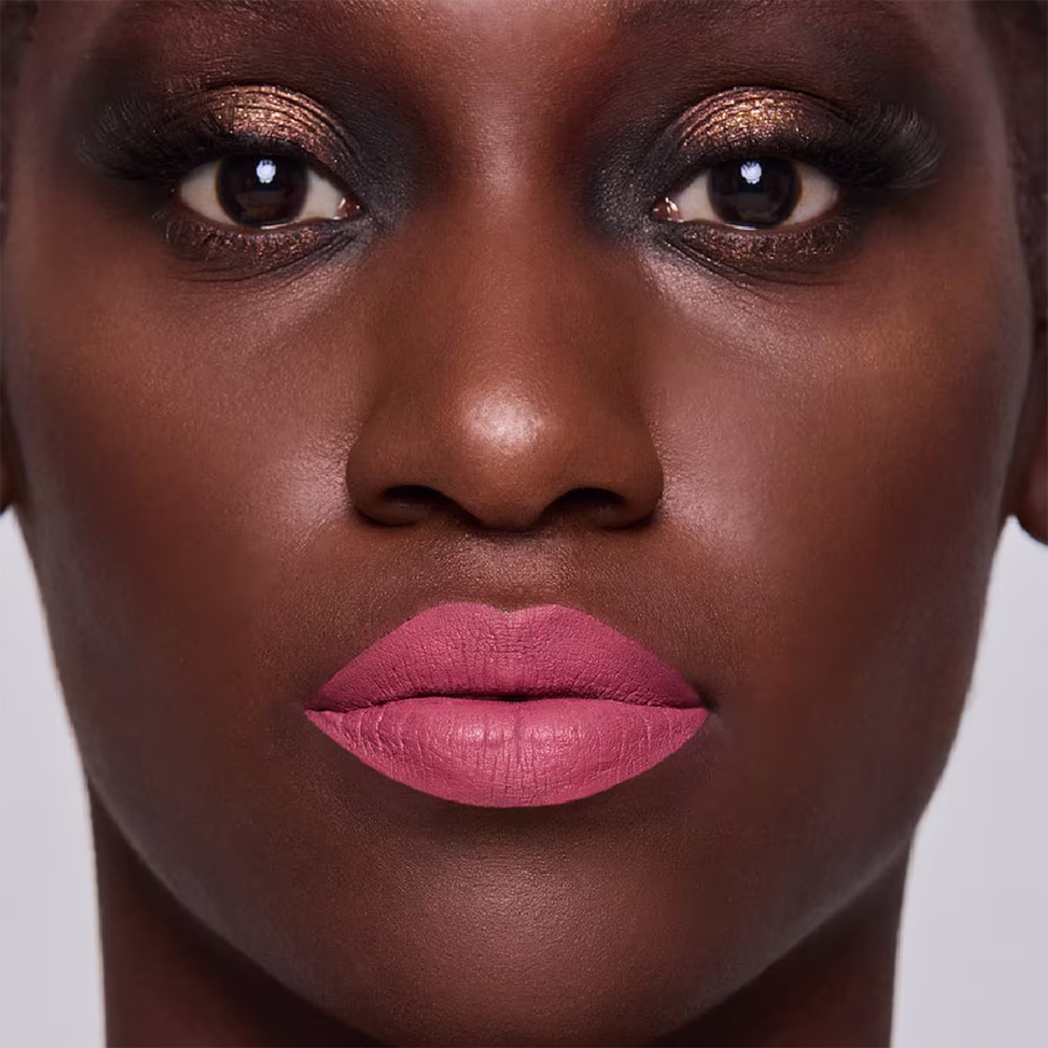 Esta imagem mostra uma mulher negra com maquiagem elaborada. A pele tem um lindo tom marrom vibrante e brilhante. Os lábios foram maquiados com um batom de cor rosa claro, realçando a boca bem delineada e cheia.