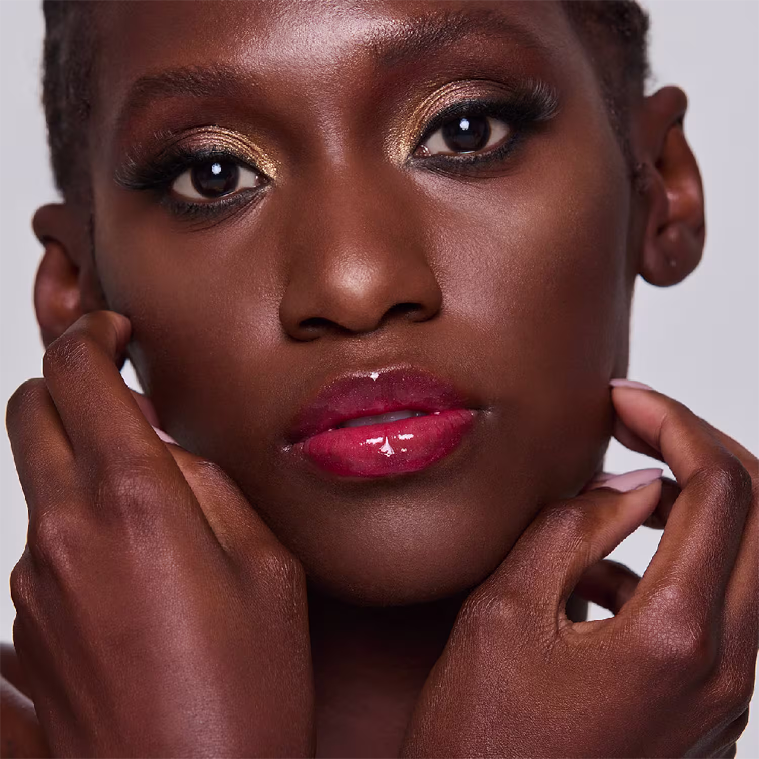 A imagem mostra uma bela modelo negra com maquiagem elaborada. Ela tem olhos intensos com sombra dourada brilhante, lábios carnudos com o gloss rosa brilhante e um lindo tom de pele bronzeado. A modelo está tocando seu rosto delicadamente com as mãos.