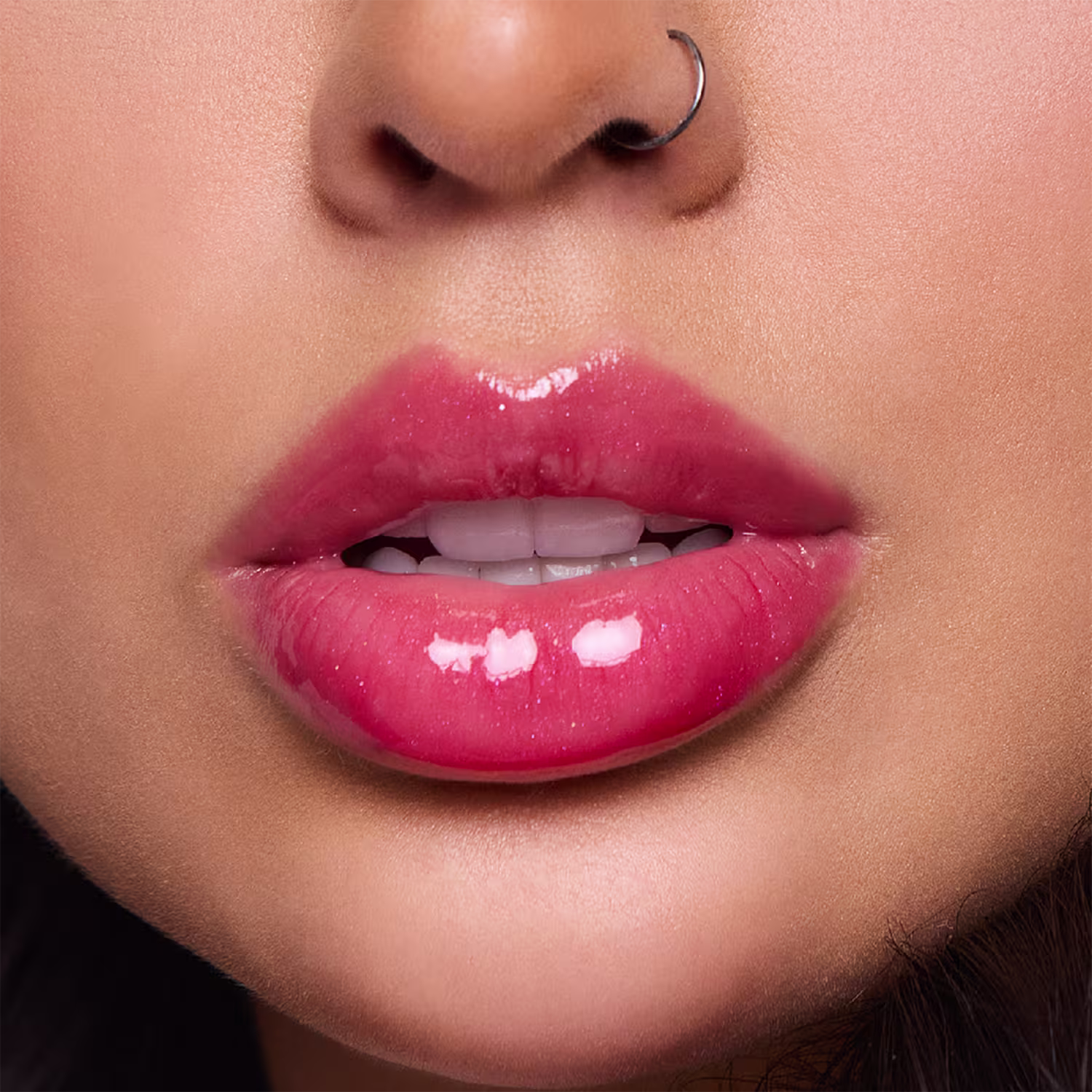 A imagem mostra um close-up dos lábios de uma mulher usando um brilho labial intenso rosado. Os lábios estão entreabertos, revelando um pouco dos dentes frontais brancos. O batom deixa os lábios com uma aparência sedosa e hidratada.