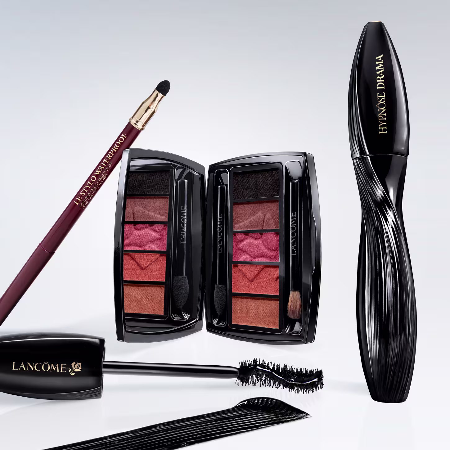 A imagem apresenta uma variedade de produtos de maquiagem da marca Lancôme. Há um lápis de olho marrom avermelhado, uma paleta de sombras com tons terrosos e rosados, um frasco preto de máscara para cílios e um aplicador de máscara de cílios.